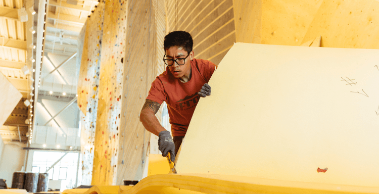 A man cuts foam for flooring in a tall, bright climbing gym