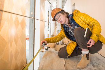 A woman with a hardhat and a measuring tape in front of a climbing wall